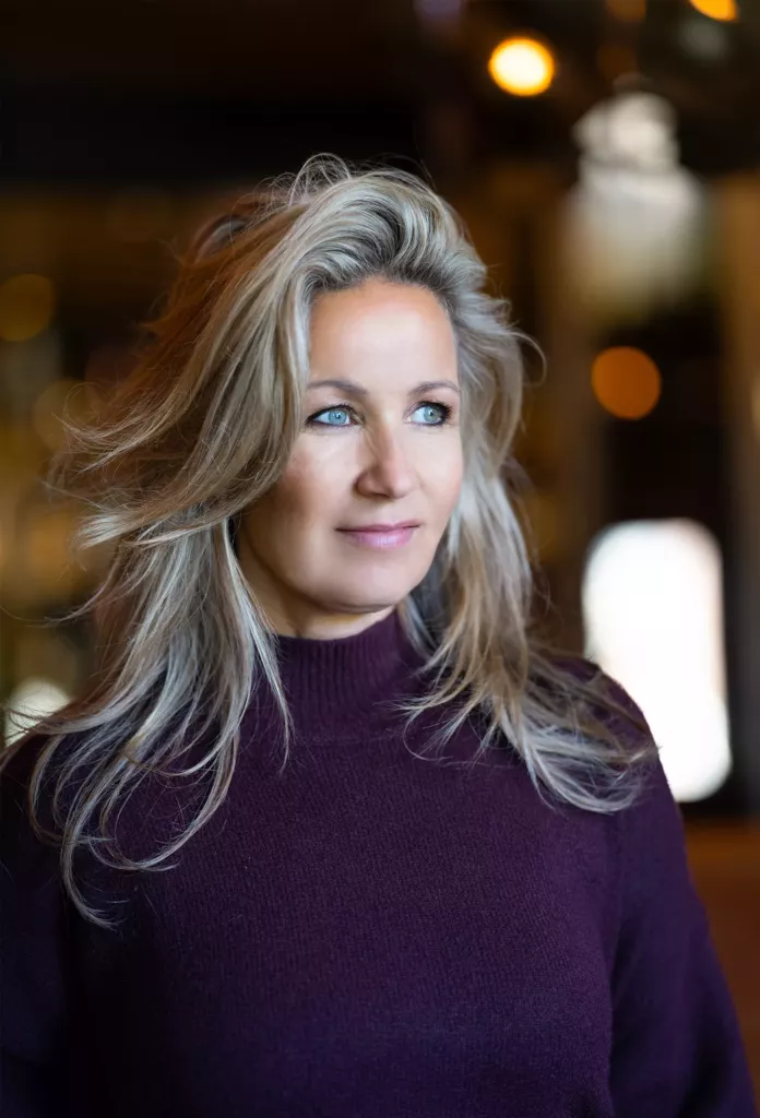 Portret van een vrouw met lang grijsblond haar in een paarse coltrui, met warme lichtjes op de achtergrond