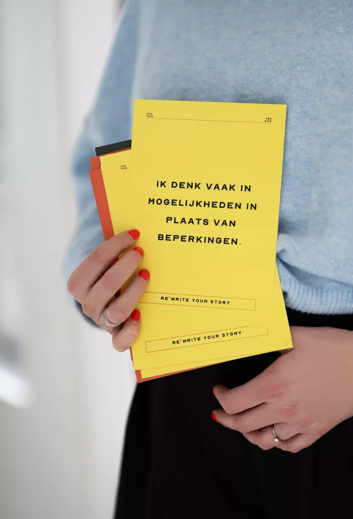 Hand met rode nagels houdt een geel boekje met de tekst Ik denk vaak in mogelijkheden in plaats van beperkingen