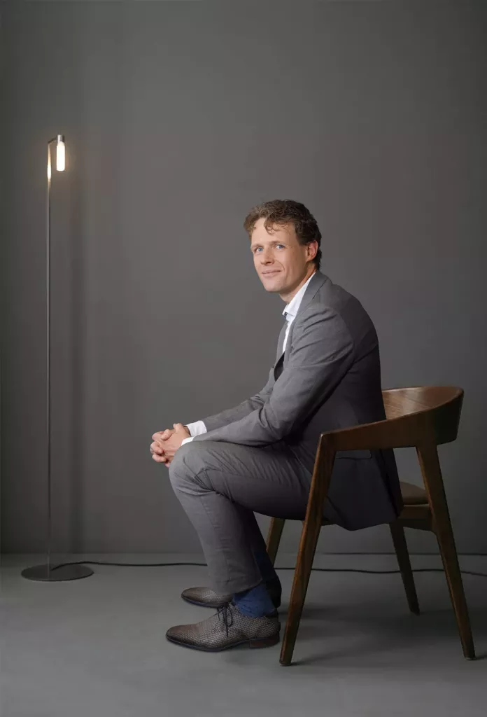 Man in een grijs pak zit op een houten stoel in een grijze studio