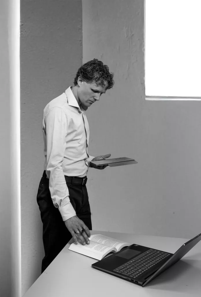 Zwart-witfoto van een man die bij een tafel met laptop in een tijdschrift bladert