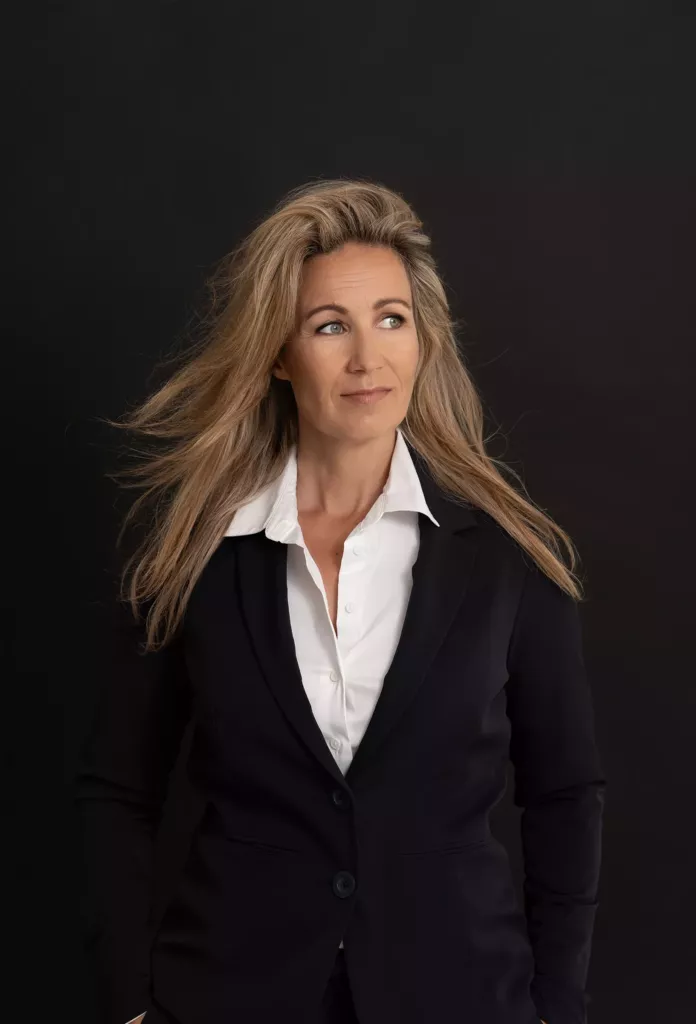 Vrouw met lang blond haar in een zwart colbert en witte blouse tegen een donkere achtergrond