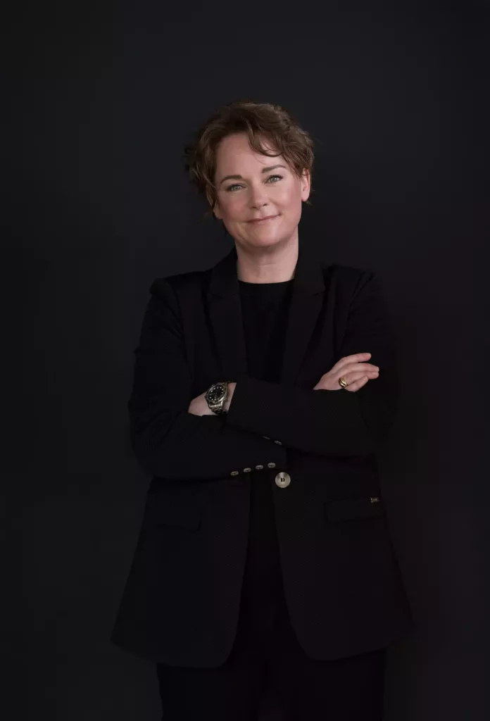 Vrouw met kort donker haar in een zwart colbert staat met gekruiste armen tegen een zwarte achtergrond