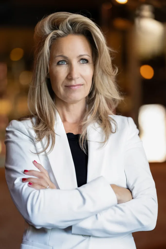 Professionele vrouw met een witte blazer en gekruiste armen, kijkt zelfverzekerd met een vage achtergrond.