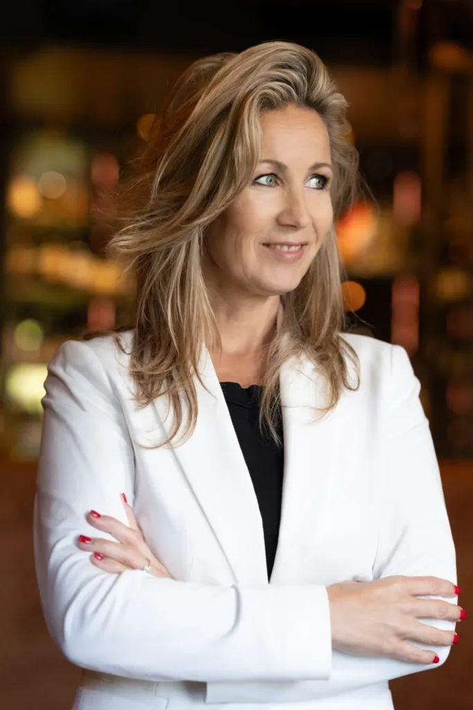 Vrouw in een witte blazer met gekruiste armen, glimlachend en kijkend naar de zijkant in een sfeervolle omgeving.