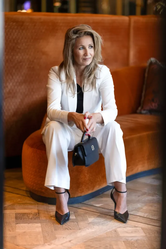 Een vrouw in een witte outfit zit op een oranje bank en houdt een zwarte handtas vast, met een ontspannen uitstraling.