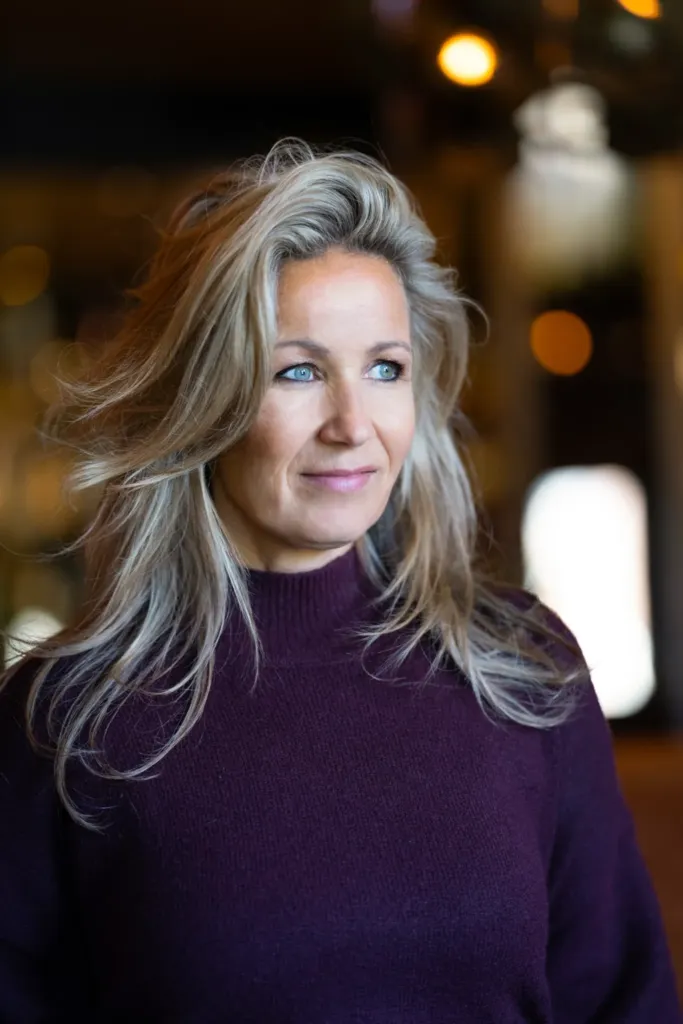 Vrouw met lang, golvend haar en blauwe ogen, gekleed in een paarse trui, kijkt met een vriendelijke uitdrukking.