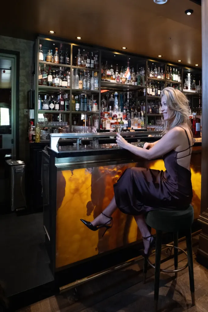 Een vrouw in een elegante jurk zit aan de bar met een cocktail, omringd door een uitgebreide selectie dranken.