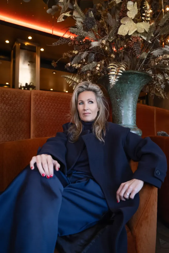 Een vrouw in een stijlvolle outfit zit ontspannen op een bank, omringd door een elegante bloemenarrangement.
