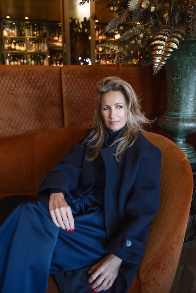 Een vrouw in een donkerblauwe outfit zit ontspannen op een oranje bank in een sfeervolle bar.