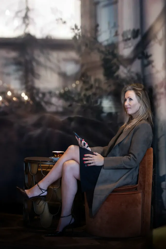 Een vrouw in een stijlvolle outfit zit ontspannen met een smartphone, terwijl ze geniet van een drankje in een chique omgeving.