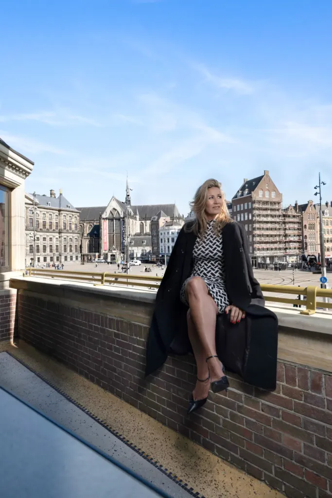 Vrouw in een zwart-witte jurk en jas, zittend op een muur met uitzicht op een druk plein en historische gebouwen.