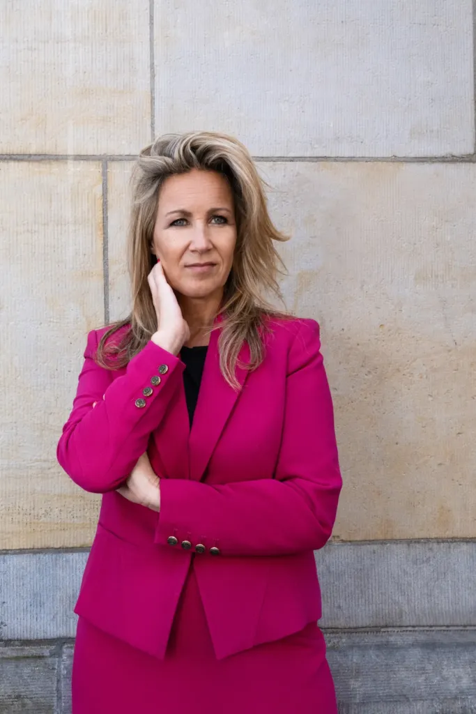 Vrouw in een fuchsia blazer staat met een zelfverzekerde houding tegen een stenen muur.