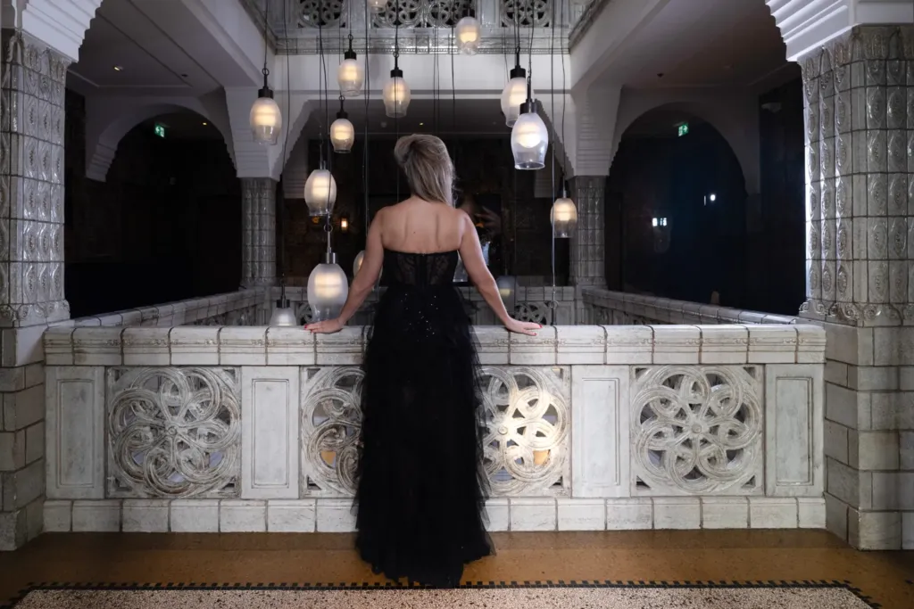 Een vrouw in een zwarte jurk staat bij een elegante balustrade, omringd door sfeervolle hanglampen.
