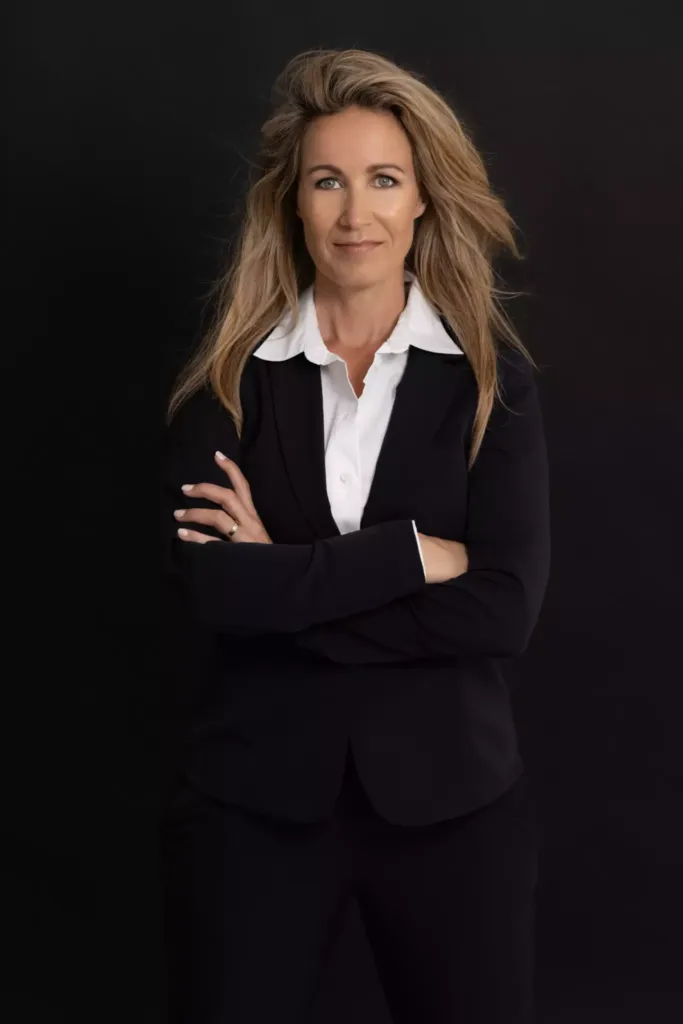 Professionele vrouw met een zelfverzekerde uitstraling, gekleed in een zwart pak met een witte blouse, tegen een zwarte achtergrond.
