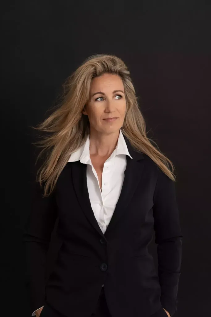 Professionele vrouw met lang, golvend haar in een zwart pak, staand voor een donkere achtergrond.