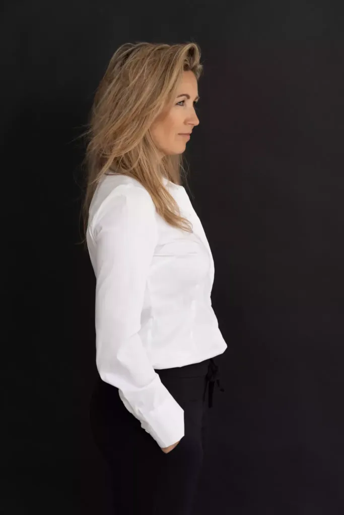 Vrouw in een witte blouse en zwarte broek, staand in profiel tegen een zwarte achtergrond.