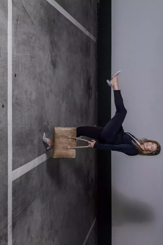 Vrouw in een parkeerplaats poseert met een tas en heft een been op, gekleed in een zwarte outfit en hakken.