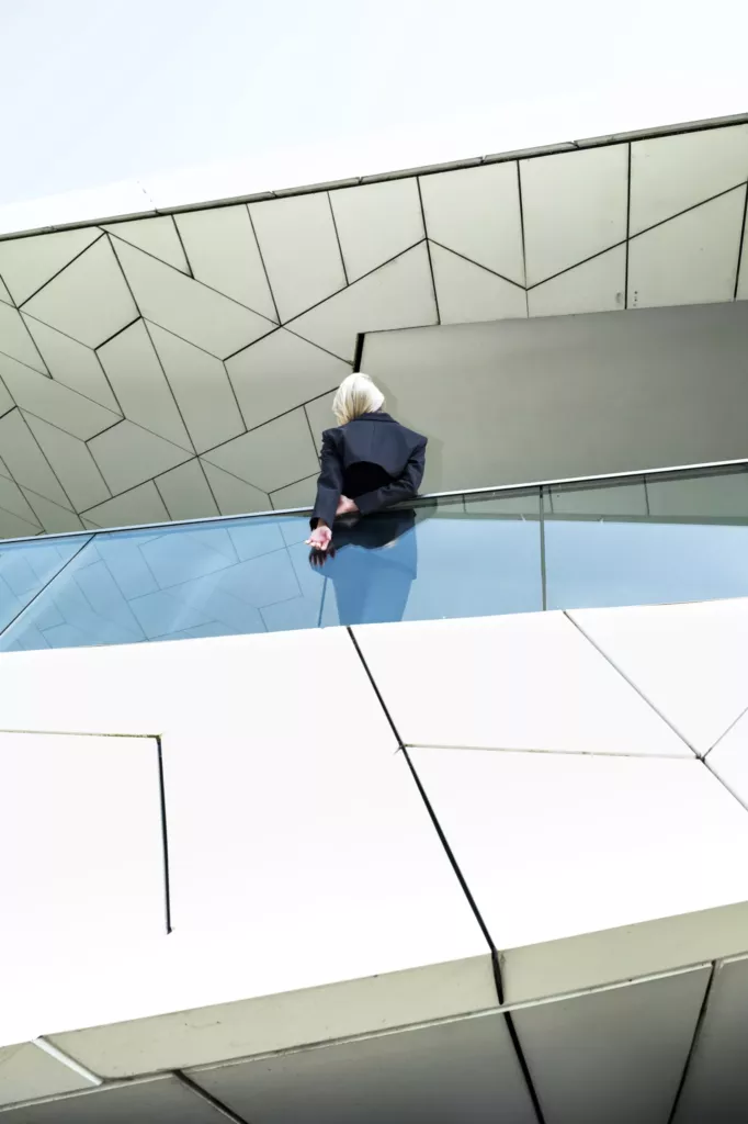 Persoon in donkere kleding zit op de glanzende, hoekige witte gevel van een modern gebouw onder een heldere lucht.