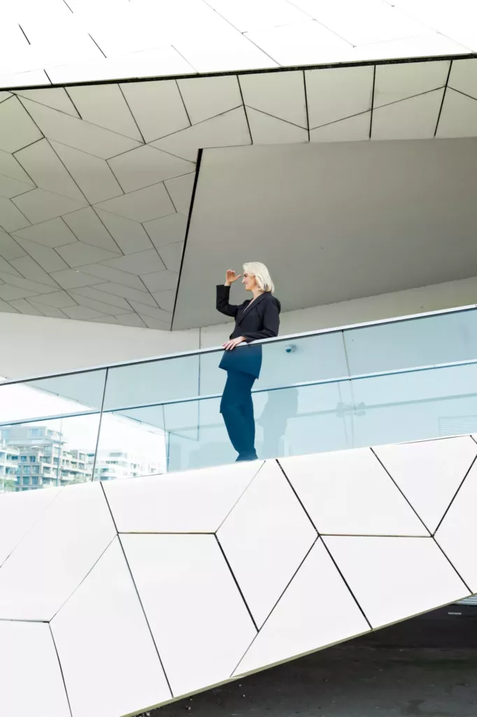 Persoon met blond haar, donkere trui en blauwe broek leunt op een glazen balustrade van een wit, hoekig gebouw.