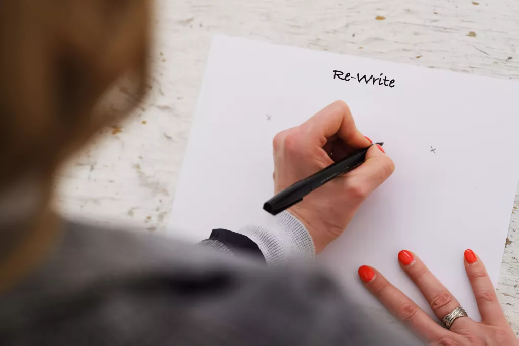 Close-up van een hand die met rode nagellak het woord 'Re-write' op papier schrijft.