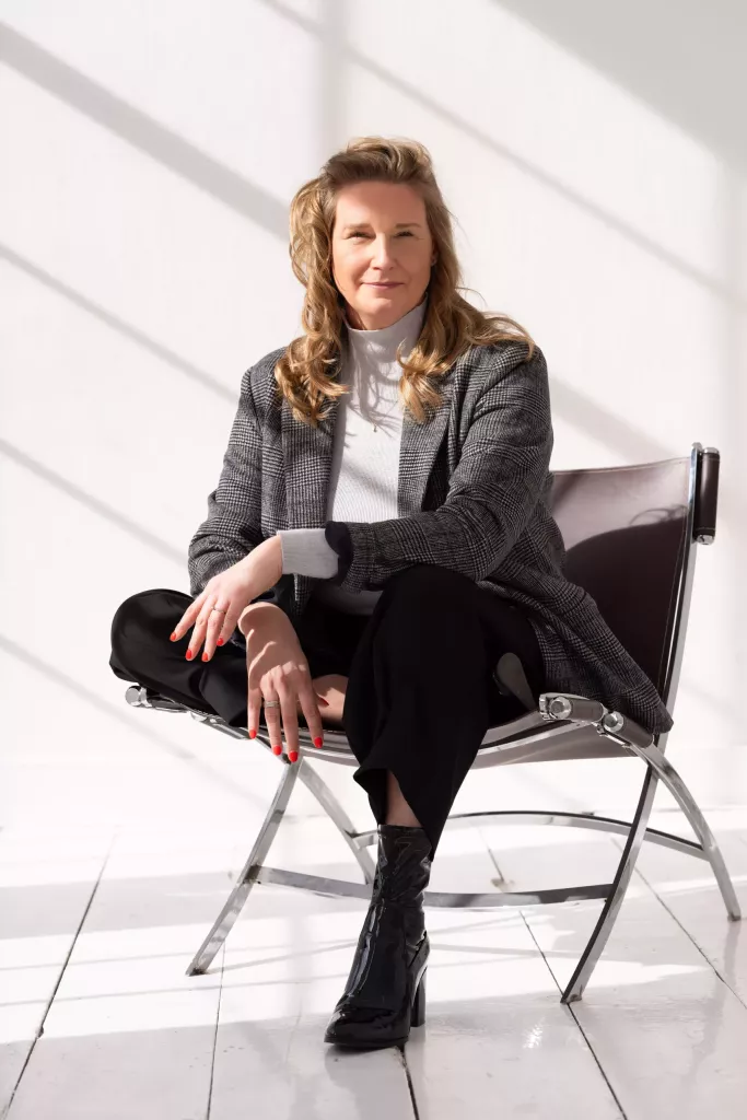 Cynthia zit glimlachend in een lounge chair van leer en chroom, met zonlicht dat strepen over de vloer werpt.
