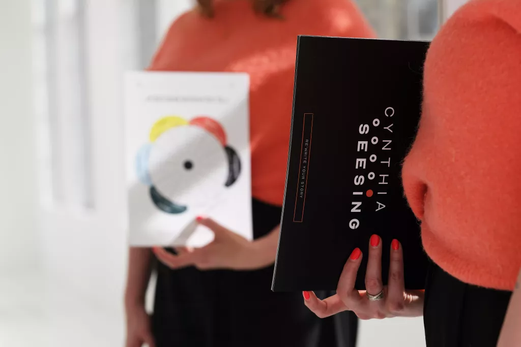 Twee handen houden een zwarte doos met opschrift Cynthia Seesing vast, naast een kaart met een kleurrijk rond motief.