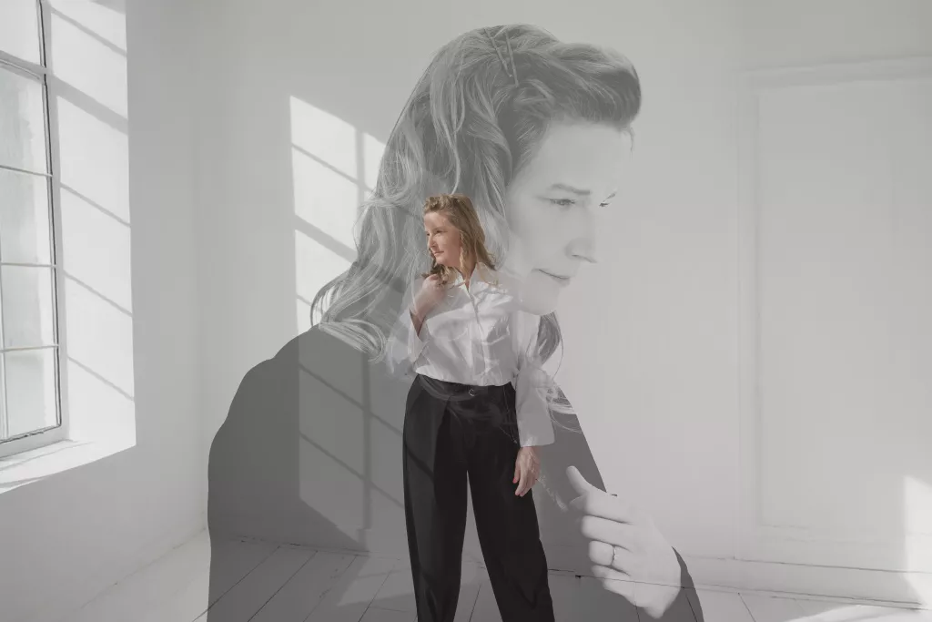Dubbelbelichting van Cynthia staand in een lichte ruimte, met een vervaagd groot portret van haar gezicht op de achtergrond.
