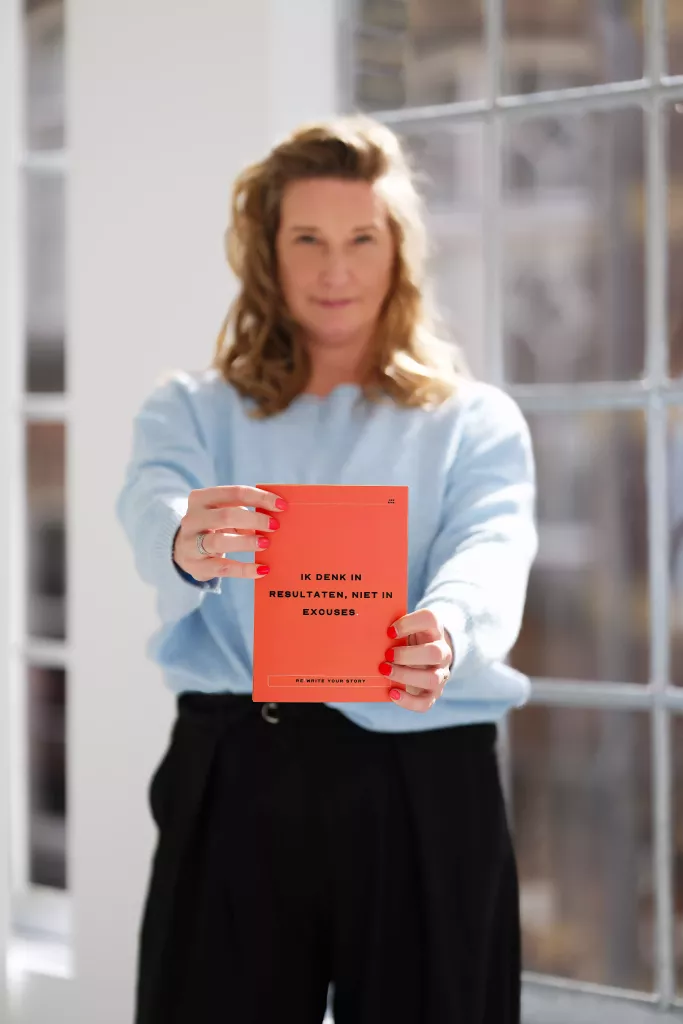 Cynthia houdt een oranje boek omhoog terwijl ze bij een raam staat, in lichtblauwe trui en zwarte broek.