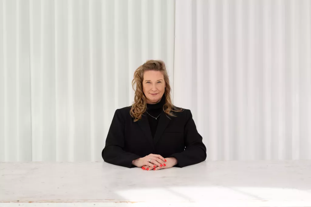 Cynthia zit in zwarte blazer met gevouwen handen aan een witte tafel, voor een geplooid wit gordijn.