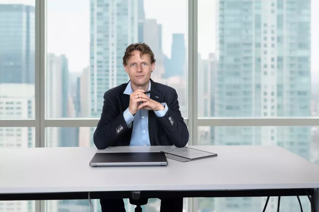 Floris Koolen aan een witte tafel met een laptop, de handen gevouwen, voor een raam met uitzicht op wolkenkrabbers