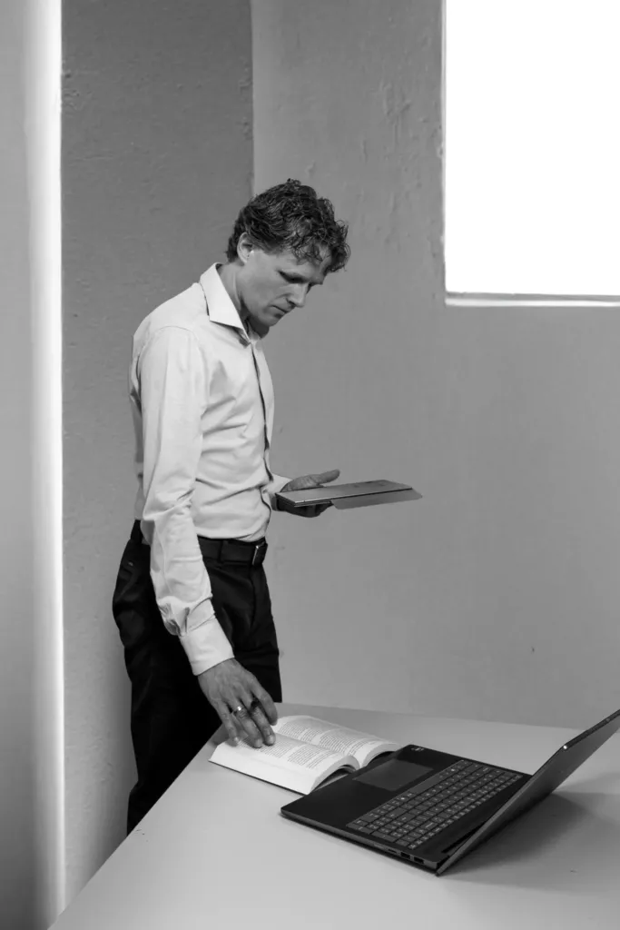 Zwart-witportret van Floris Koolen, gebogen over een tafel met een laptop terwijl hij naar een tablet kijkt