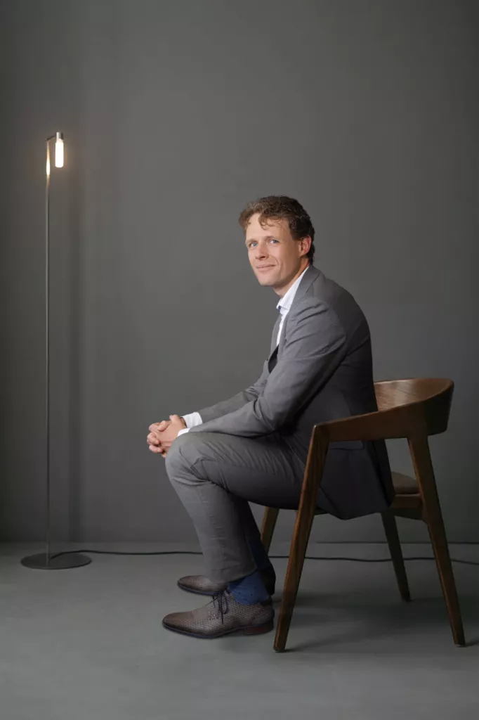 Floris Koolen in een grijs pak, zittend op een houten stoel tegen een grijze muur met een staande lamp