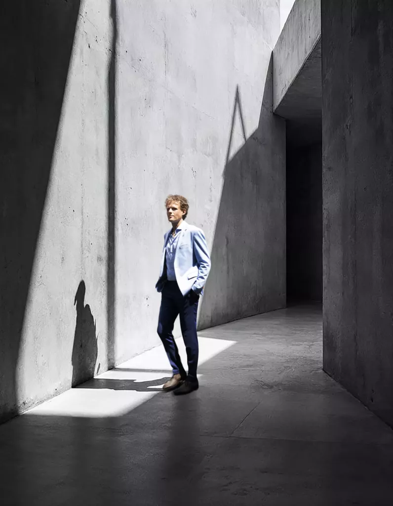 Floris Koolen in een lichtblauw jasje, staand in een betonnen ruimte met scherpe schaduwen en een streep zonlicht