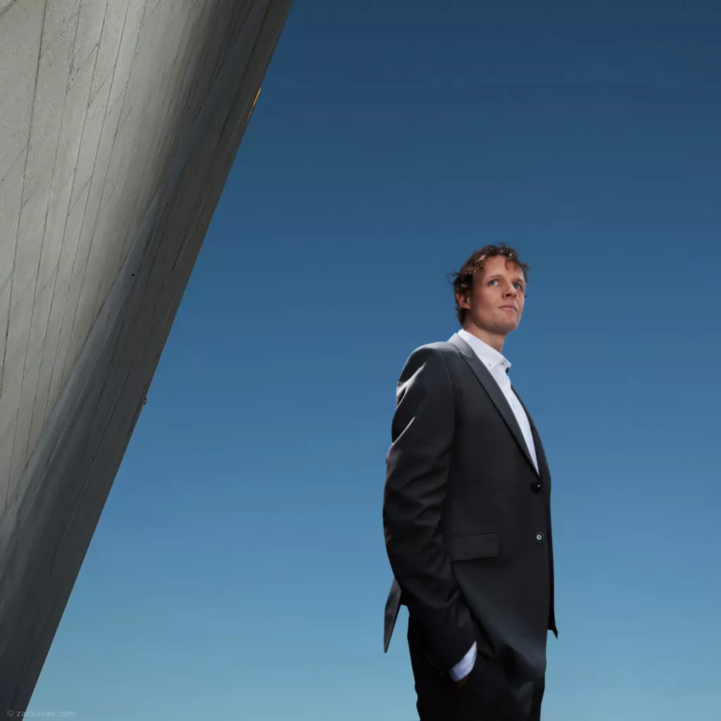 Floris Koolen in een donker pak, van onderaf gefotografeerd tegen een diepblauwe lucht naast een betonnen muur