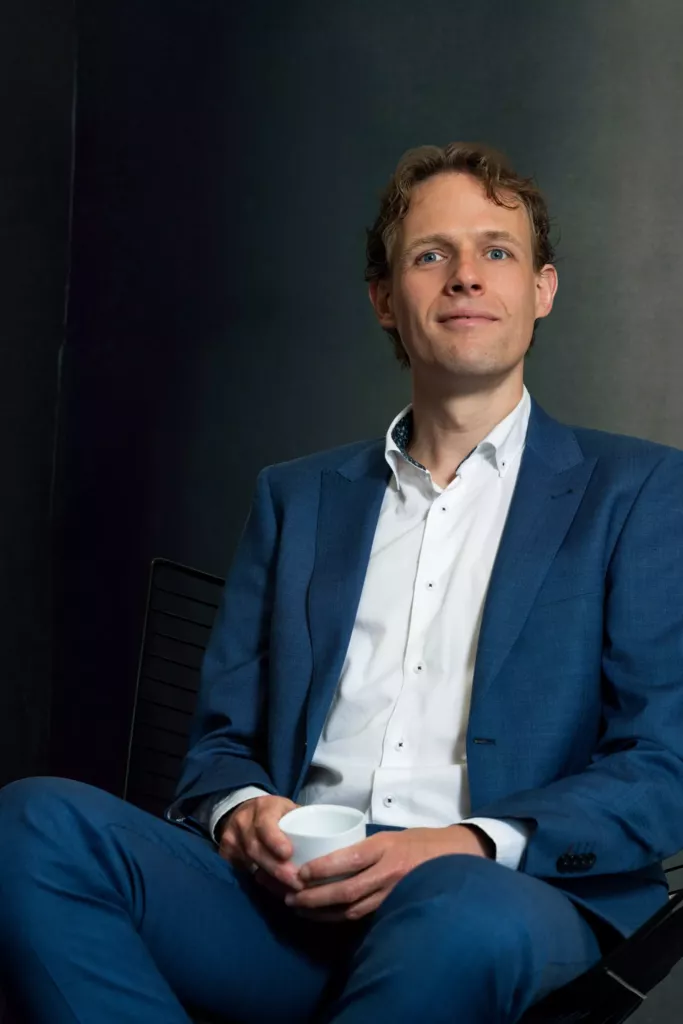 Floris Koolen in een blauw pak en wit overhemd, zittend met een kopje in zijn handen tegen een donkere achtergrond