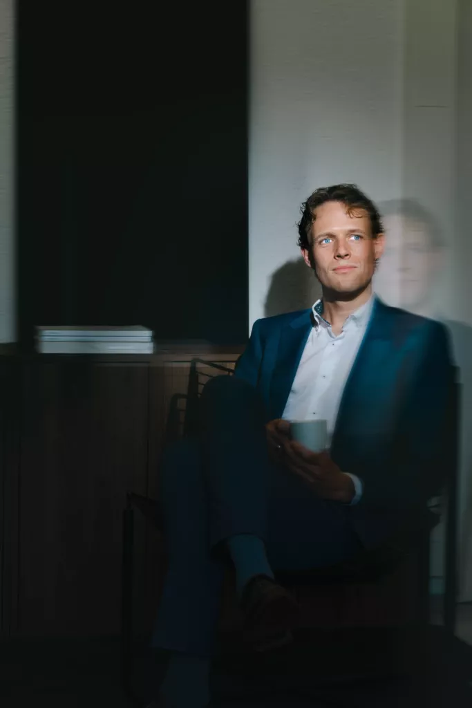 Floris Koolen in een donker pak, zittend in een schemerige ruimte met een kopje in zijn hand