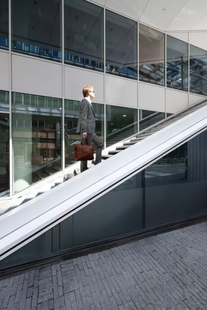 Floris Koolen loopt met een bruine tas langs een roltrap van een modern gebouw met een glazen gevel