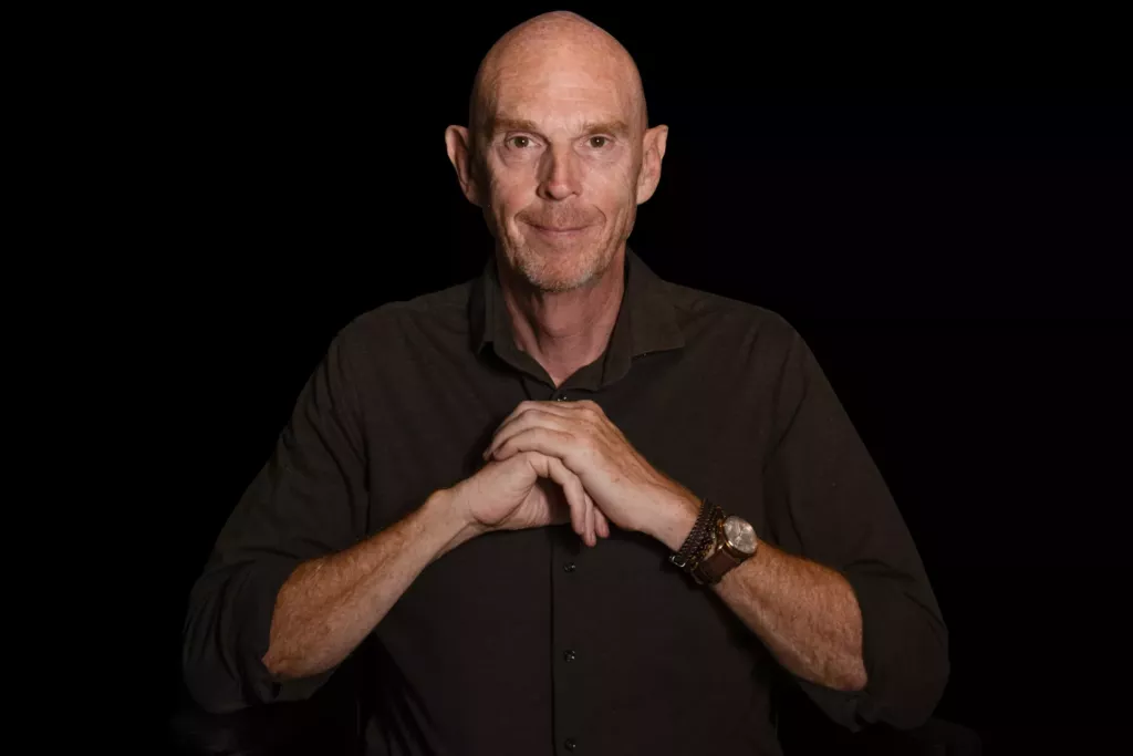 Peter, een kale man in donkere kleding, houdt zijn handen voor zijn borst tegen een zwarte achtergrond.