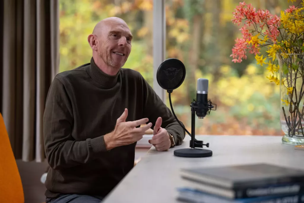 Peter zit aan een tafel met een microfoon en praat, met herfstbladeren achter het raam en boeken op de voorgrond.