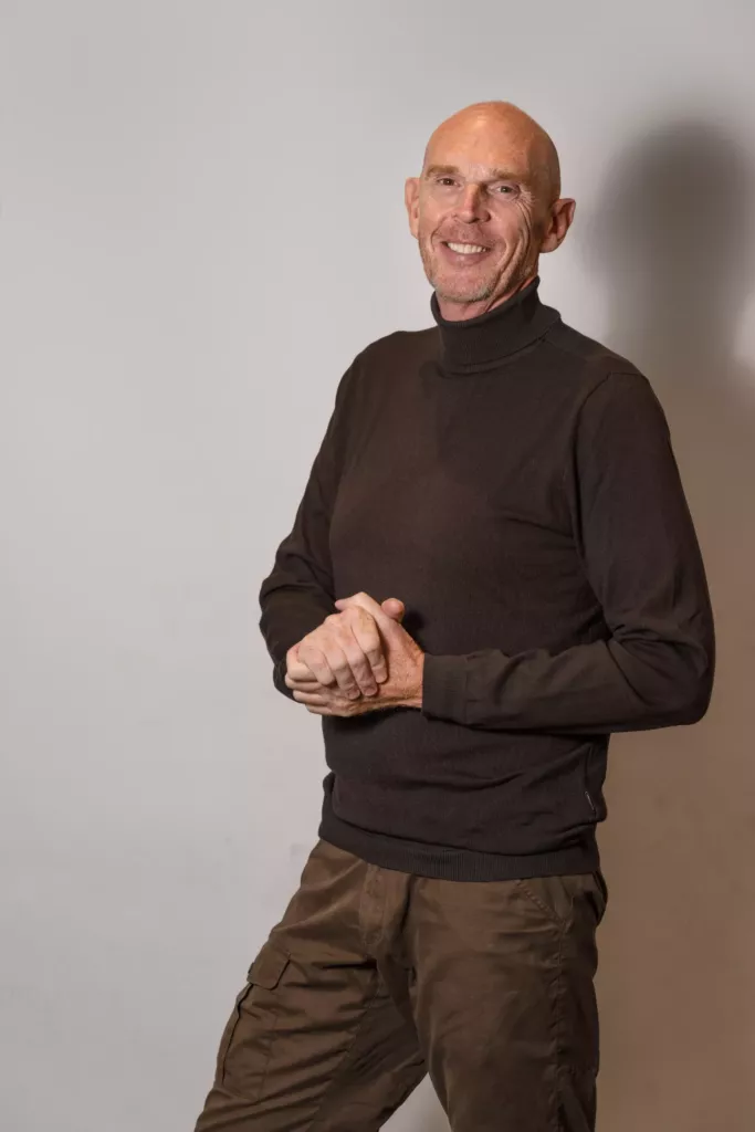 Peter glimlacht ontspannen in een coltrui tegen een grijze achtergrond.