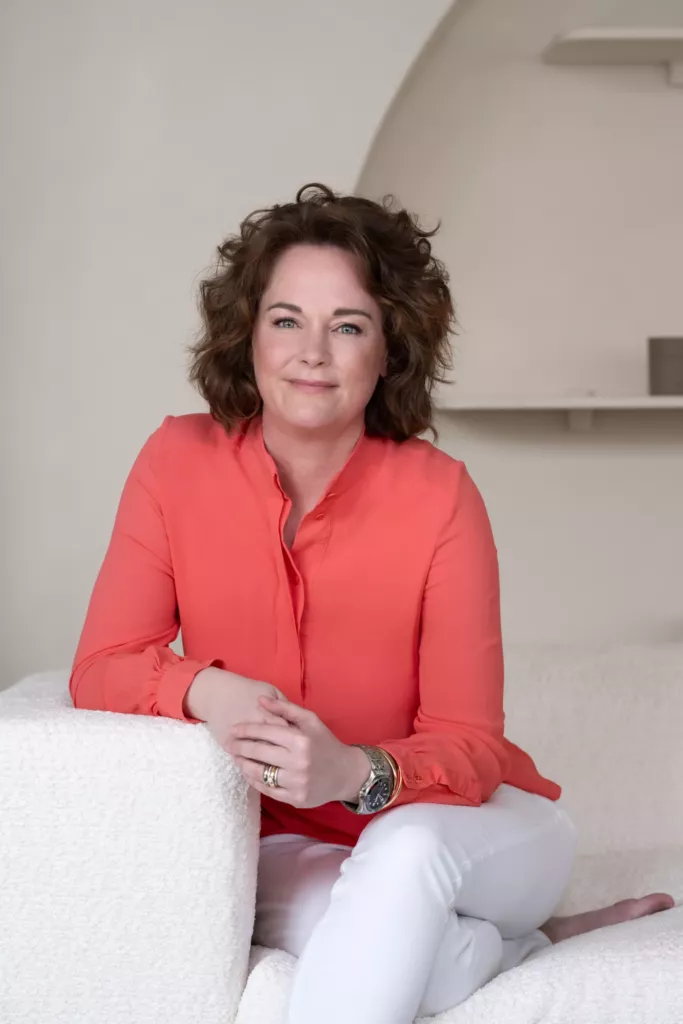 Thera zit ontspannen op een witte bank in een koraalkleurige blouse.