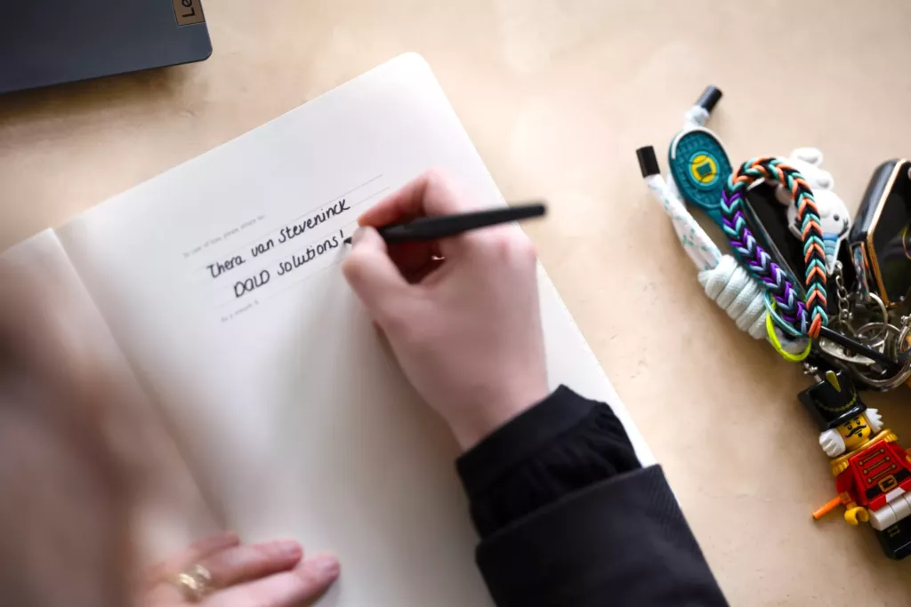 Thera schrijft met een zwarte stift op papier op een houten bureau, met kleurrijke kleine voorwerpen ernaast.