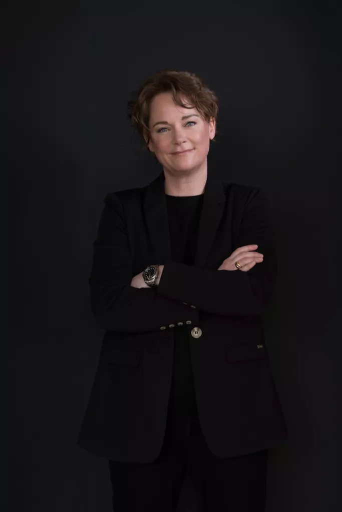 Thera in donkere kleding met de armen over elkaar tegen een donkere achtergrond.
