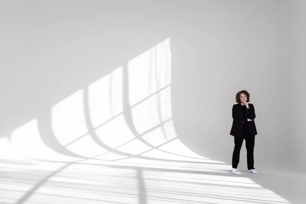 Thera staat klein in donkere kleding in een grote witte ruimte met lange schaduwen van ramen op muur en vloer.
